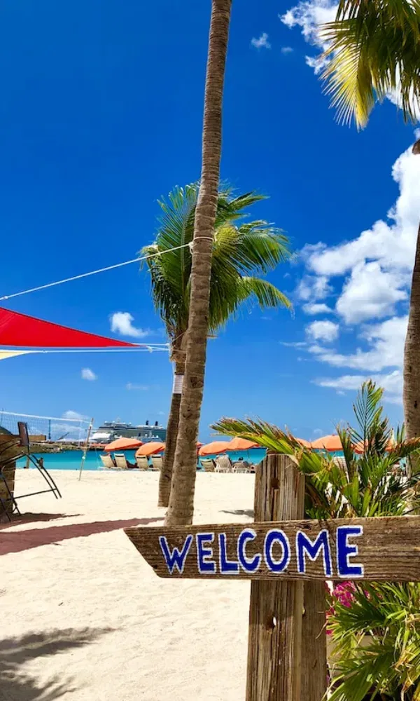 Strandkulisse mit einem handgemalten „Welcome“-Schild, tropischer Vegetation, Meerblick und einem Schiff in der Ferne.