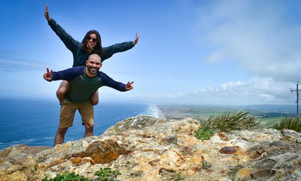 Zwei Wanderfreunde feiern ihren Gipfelerfolg mit einem verspielten Foto vor atemberaubender Aussicht auf Küste, Wellen und Himmel.