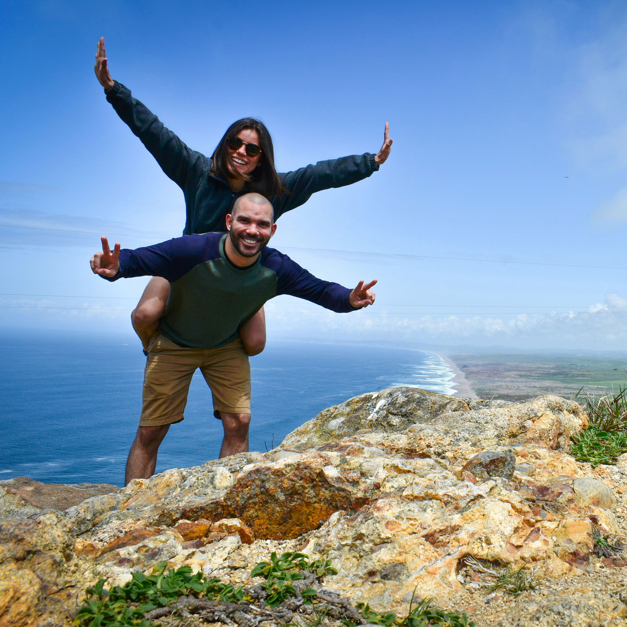 Zwei Reisende feiern ihre Aussicht auf die weite Küste von einem Felsvorsprung aus – die Frau sitzt mit ausgebreiteten Armen auf den Schultern ihres Partners.