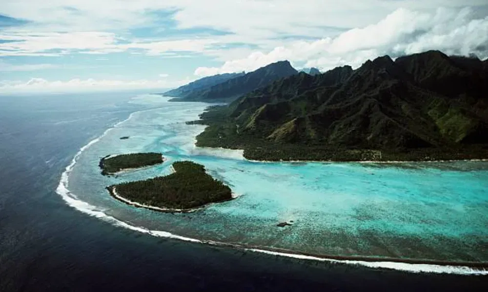 Luftbild einer pazifischen Inselküste mit türkisfarbenem Wasser, Korallenriffen und bewaldeten Landzungen entlang der Berge.