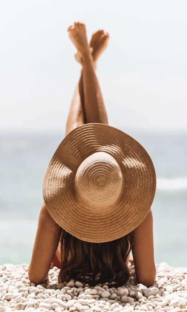 Rückenansicht einer Frau, die entspannt auf einem steinigen Strand liegt – Beine überkreuzt, Strohhut schützt vor der Sonne, Meer im Hintergrund.