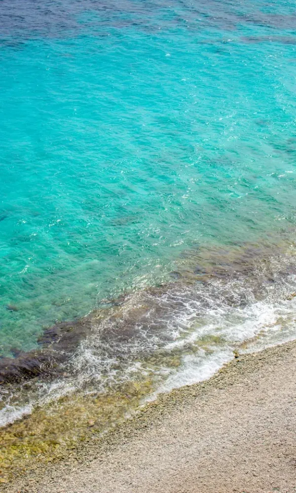 Blick auf einen Kiesstrand mit leichtem Wellengang und türkisblauem Meer, das sich zum Horizont hin verdunkelt.