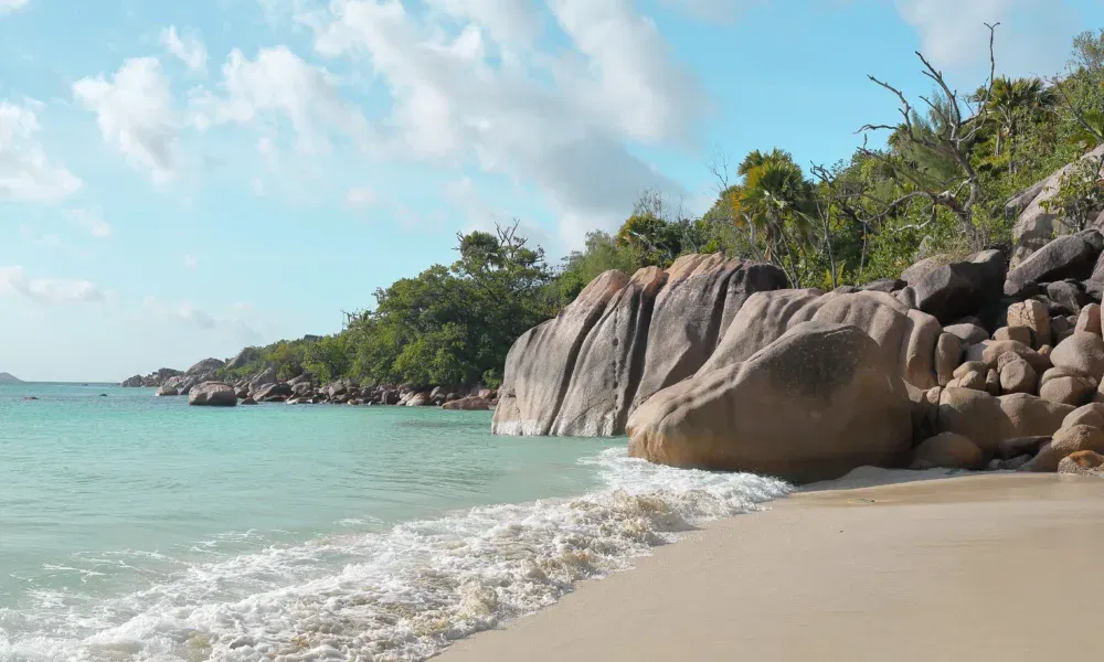 Ruhiger Sandstrand mit türkisfarbenem Wasser, umrahmt von runden Granitfelsen und tropischer Vegetation.