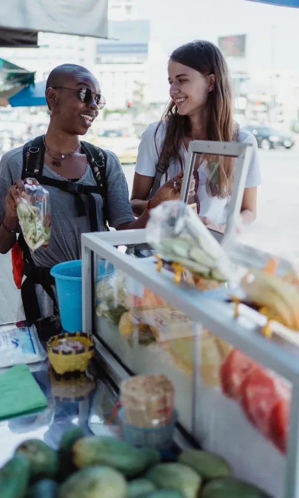 Marktszene mit zwei Touristinnen, die an einem Street-Food-Stand frisches Obst begutachten und sich fröhlich unterhalten.