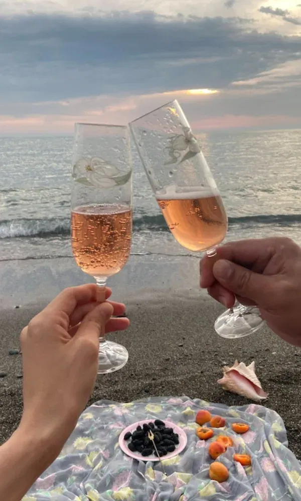 Feierlicher Moment am Meer: Drei Hände mit Gläsern Rosé-Sekt stoßen bei einem Picknick direkt am Wasser auf den Abend an.