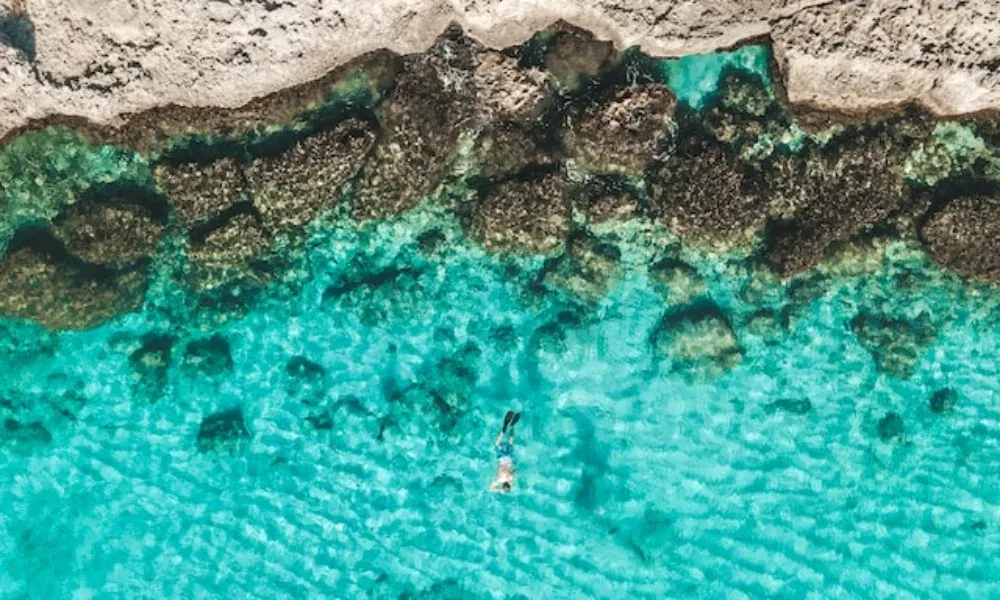 Kristallklares Wasser und eine einsame Schnorchlerin an einer felsigen Küstenlinie – tropische Idylle aus der Vogelperspektive.