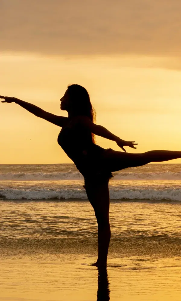 Tänzerin am Meer bei Sonnenuntergang, in eleganter Pose auf einem Bein balancierend im nassen Sand.