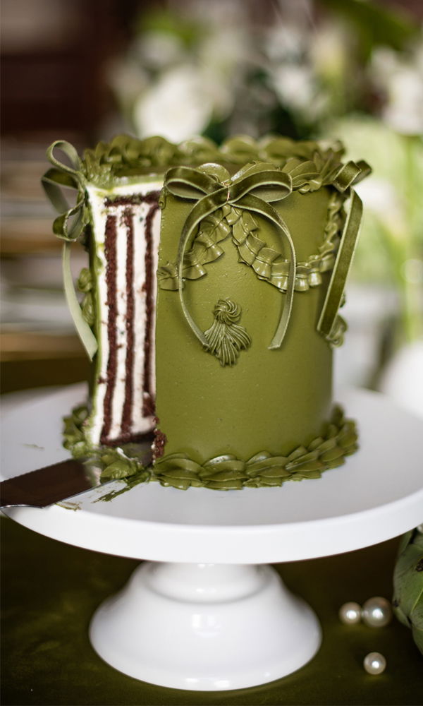 Diese einzigartige Hochzeitstorte in sattem Grün erinnert an eine luxuriöse Geschenkbox mit handgefertigten Fondant-Schleifen und Bordüren. Der offene Schnitt gibt den Blick auf kontrastreiche Schokoladen- und Vanilleböden frei. Die Torte ruht auf einem schlichten, weißen Kuchensockel, während der Hintergrund in sanften Grüntönen gehalten ist.