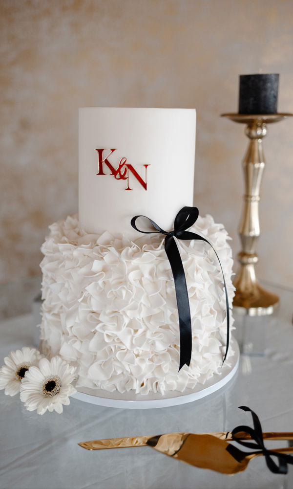 Elegante Hochzeitstorte in Weiß mit raffinierter Struktur. Der untere Teil ist von zarten, voluminösen Zuckerblüten umhüllt, während der obere minimalistisch bleibt und mit roten Monogramm-Buchstaben sowie einer schwarzen Schleife dekoriert ist. Eine goldene Tortenschaufel und dezente Blumendetails ergänzen die Szene.