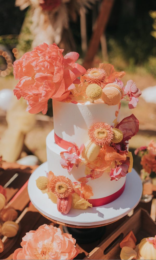 Zweistöckige Hochzeitstorte mit strahlend weißen Fondantschichten, umrahmt von kräftigen Farben. Opulente Pfingstrosen, filigrane Trockenblumen und Macarons in Orange, Gelb und Pink verleihen der Torte eine dynamische, festliche Ausstrahlung. Die feinen Bänder in kräftigem Rot setzen einen harmonischen Akzent. Das sonnige Outdoor-Setting verstärkt den sommerlichen Flair der Szenerie.