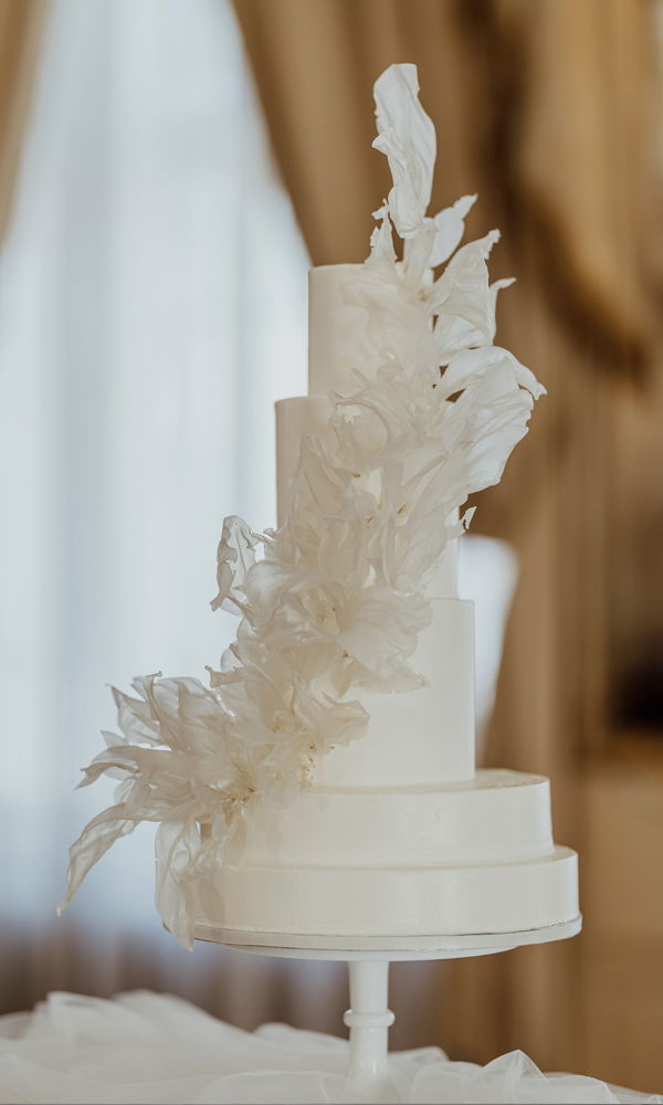 Extravagante Hochzeitstorte in minimalistischem Weiß mit vier Etagen. Die dekorativen Elemente aus feinem Reispapier verlaufen verspielt über die gesamte Torte und erzeugen eine dynamische, fast schwebende Wirkung. Der luxuriöse Hintergrund mit goldenem Lichtspiel betont die edle Ästhetik.