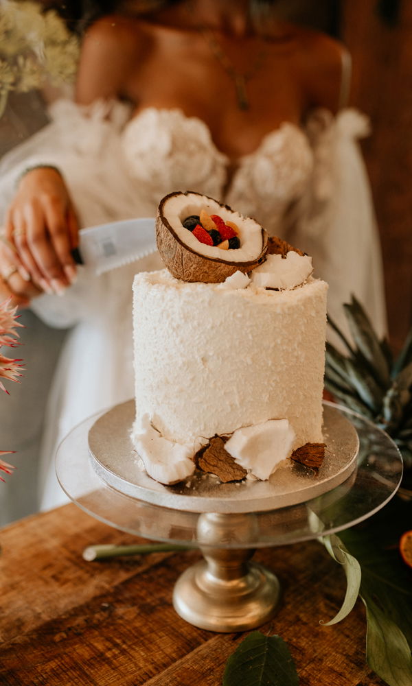 Minimalistische, aber wirkungsvolle Kokostorte mit tropischem Flair. Die Textur aus feinen Kokosraspeln harmoniert mit der natürlichen Dekoration aus Kokosnussschalen und Beeren. Die Torte steht auf einer goldenen Kuchenplatte, während eine elegant gekleidete Braut sich darauf vorbereitet, den ersten Schnitt zu setzen.