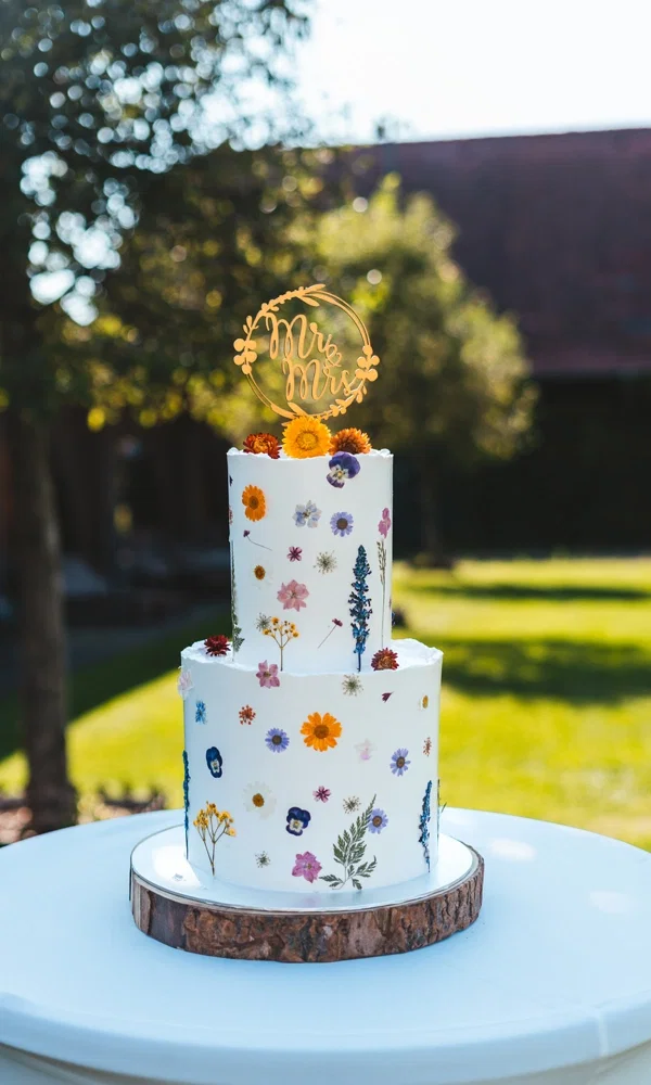 Einzigartige Hochzeitstorte mit botanischem Design: Zwei elegante Fondantschichten sind mit gepressten Blüten in verschiedenen Formen und Farben dekoriert, wodurch die Torte wie ein kunstvolles Blumenarrangement wirkt. Das goldene „Mr & Mrs“-Topper-Detail fügt einen stilvollen Akzent hinzu. Präsentiert auf einer Baumscheibe und umgeben von Natur, spiegelt die Torte eine harmonische Verbindung zwischen Eleganz und rustikalem Charme wider.