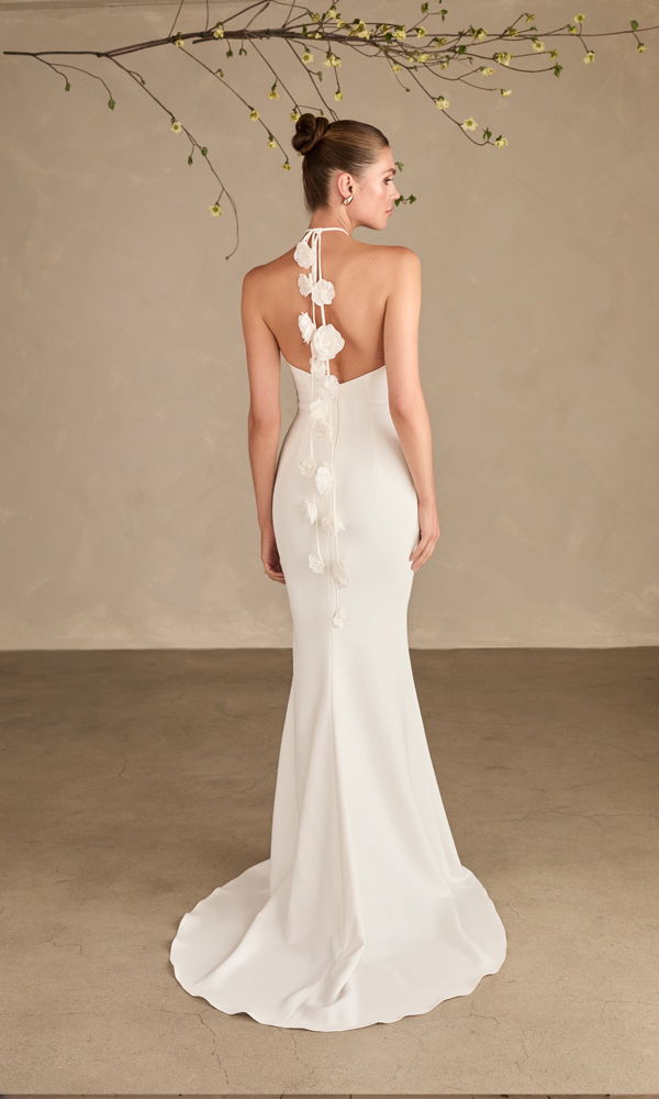 Schlichtes Hochzeitskleid mit einzigartigem Blumenband-Detail am Rücken, präsentiert in einer puristischen Umgebung.