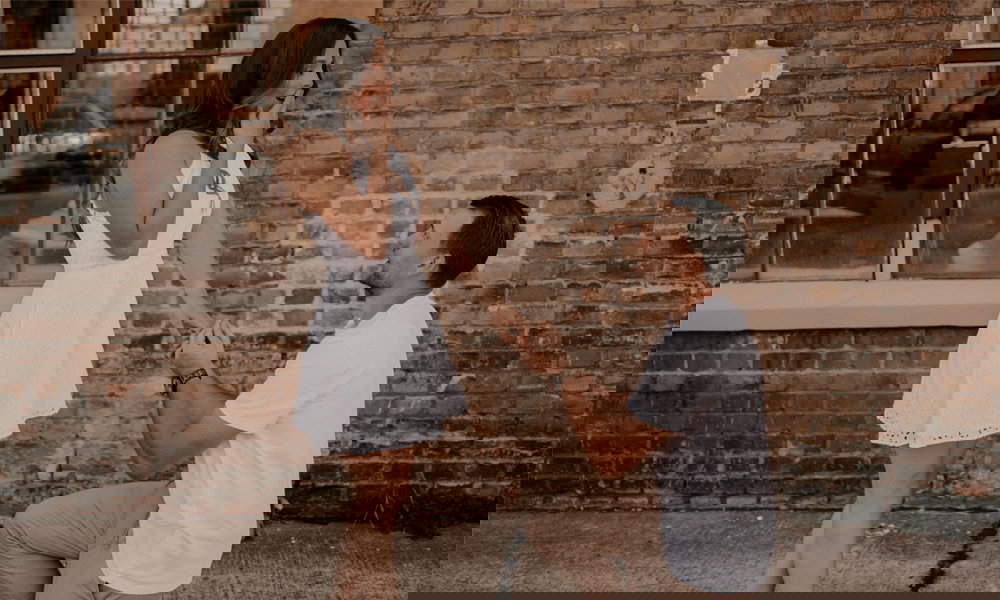 Ein überraschender Moment: Ein Mann macht seiner Partnerin einen Heiratsantrag vor einer rustikalen Wand, sie hält sich vor Freude die Hand vor den Mund.