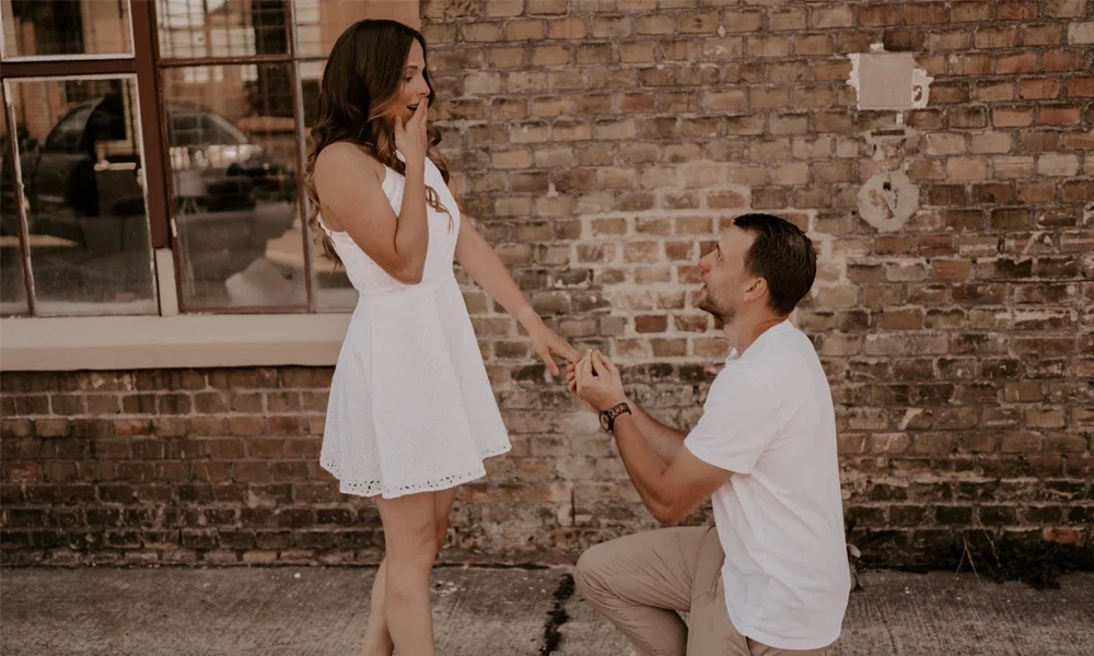 Ein überraschender Moment: Ein Mann macht seiner Partnerin einen Heiratsantrag vor einer rustikalen Wand, sie hält sich vor Freude die Hand vor den Mund.