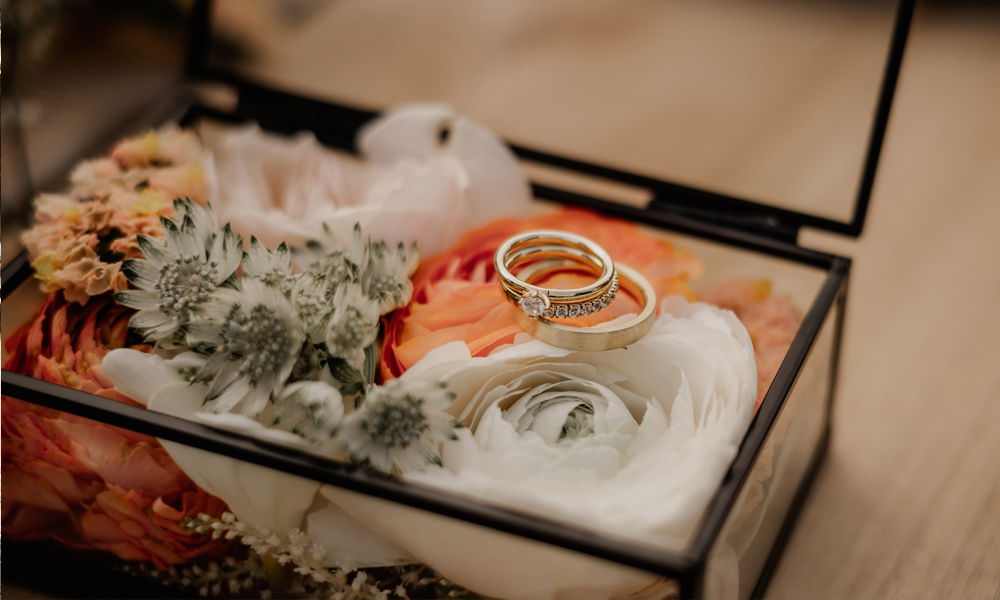 Ein Paar goldene Trauringe, darunter ein mit Diamanten verzierter Ring, ruht in einer transparenten Box auf einer Anordnung aus hellen und bunten Blumen.