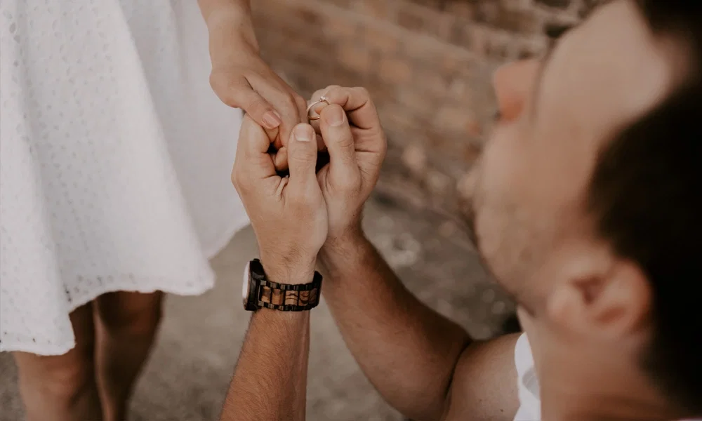 Ein Mann kniet und hält die Hand seiner Partnerin, während er ihr zärtlich einen Verlobungsring an den Finger steckt, ein Symbol für Liebe und Engagement.