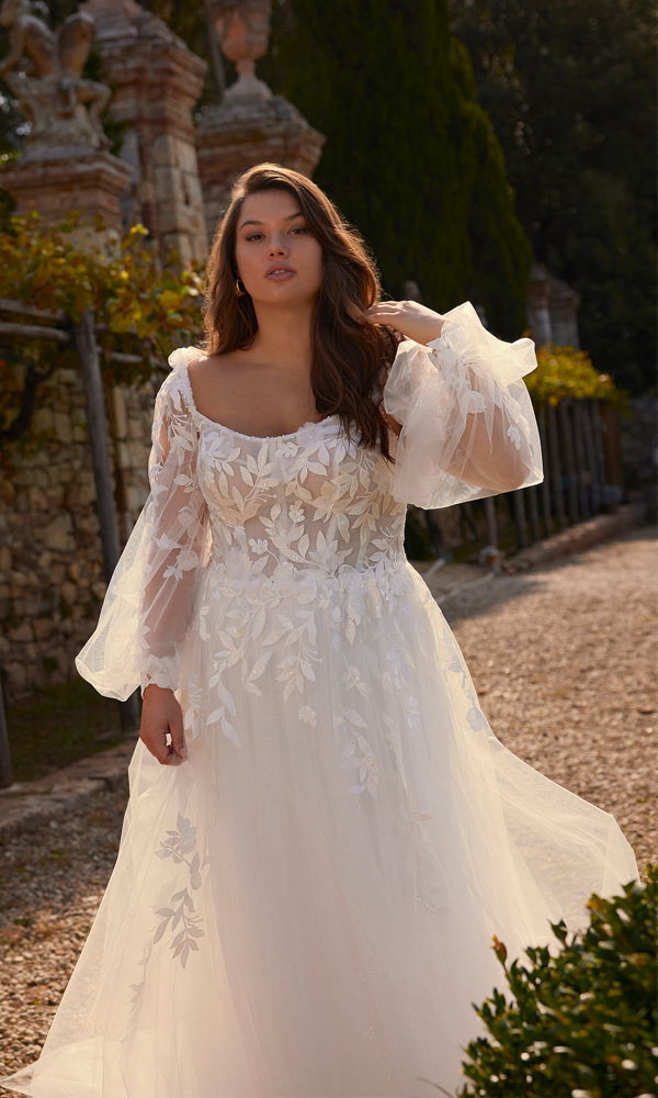 Elegantes Brautkleid mit tiefem Ausschnitt, floralen Spitzenapplikationen und transparenten Tüllärmeln. Die Kombination aus verspielter Leichtigkeit und edler Spitze sorgt für einen einzigartigen Look.