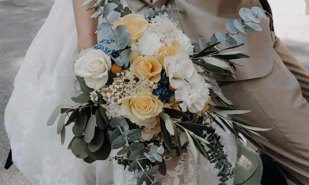 Detailaufnahme eines sommerlichen Brautstraußes mit gelben und weißen Rosen, kleinen Blüten und grünem Eukalyptus. Die Braut trägt ein Spitzenkleid, während der Bräutigam in Beige neben ihr sitzt.