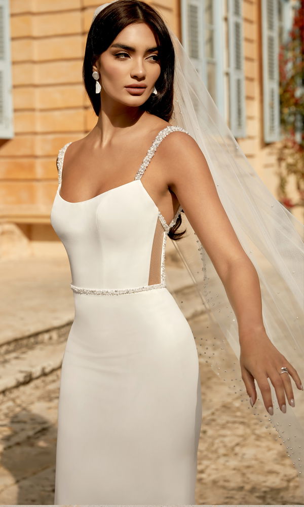 Zeitloses Brautkleid mit dezenten Details und fließender Silhouette, ergänzt durch einen langen, perlenbesetzten Schleier in einer mediterranen Kulisse.