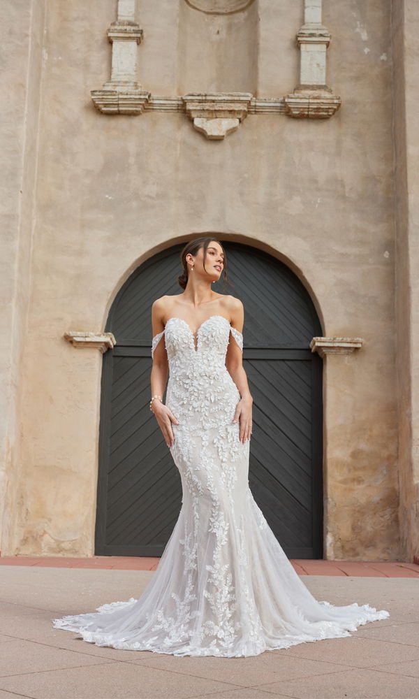 Elegantes schulterfreies Brautkleid mit floralen Applikationen und langer Schleppe, abgebildet vor einer rustikalen Steinwand mit Bogentür.
