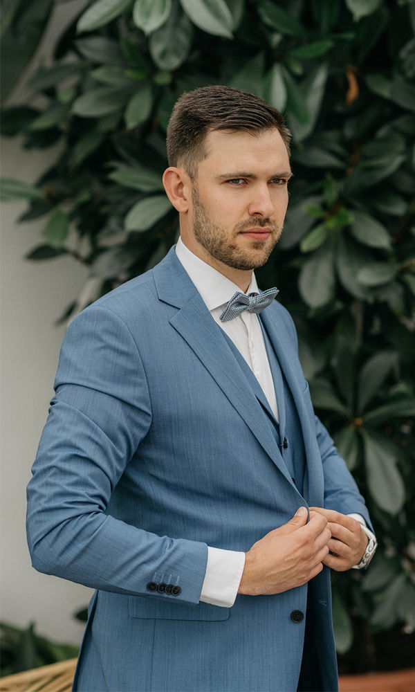 Ein stilvoll gekleideter Mann in einem blauen Anzug mit Fliege posiert vor einer Wand mit üppigen Pflanzen und sieht selbstbewusst aus.