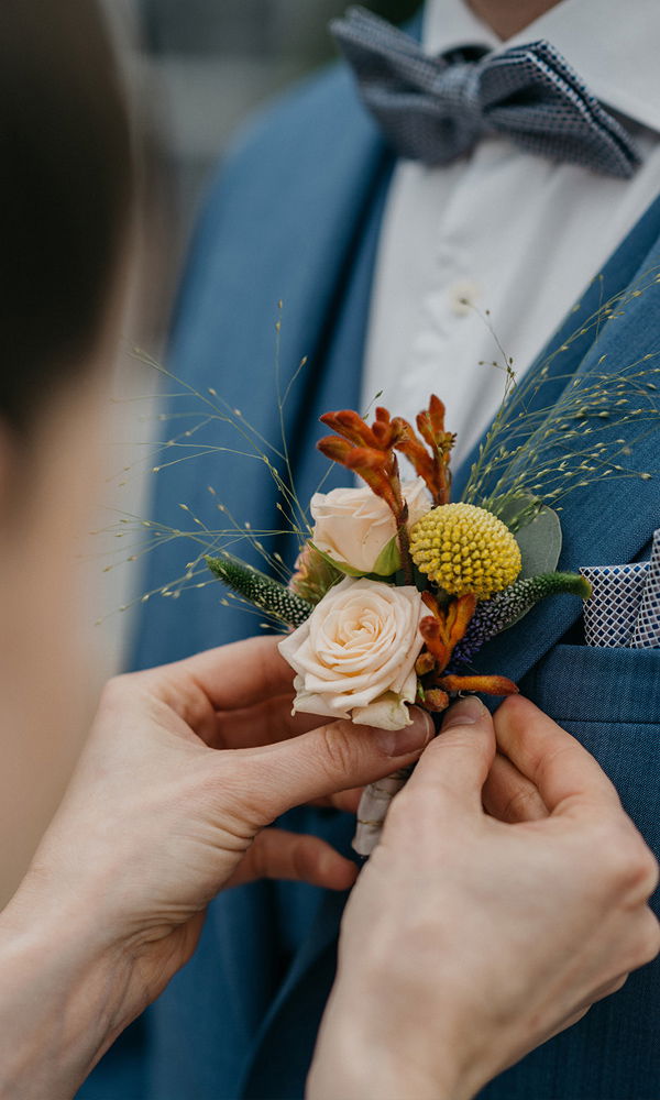 Detailaufnahme von einer Hand, die eine Boutonniere mit Rosen, gelben und orangenen Blumen sowie dekorativen Gräsern am Anzugkragen fixiert.