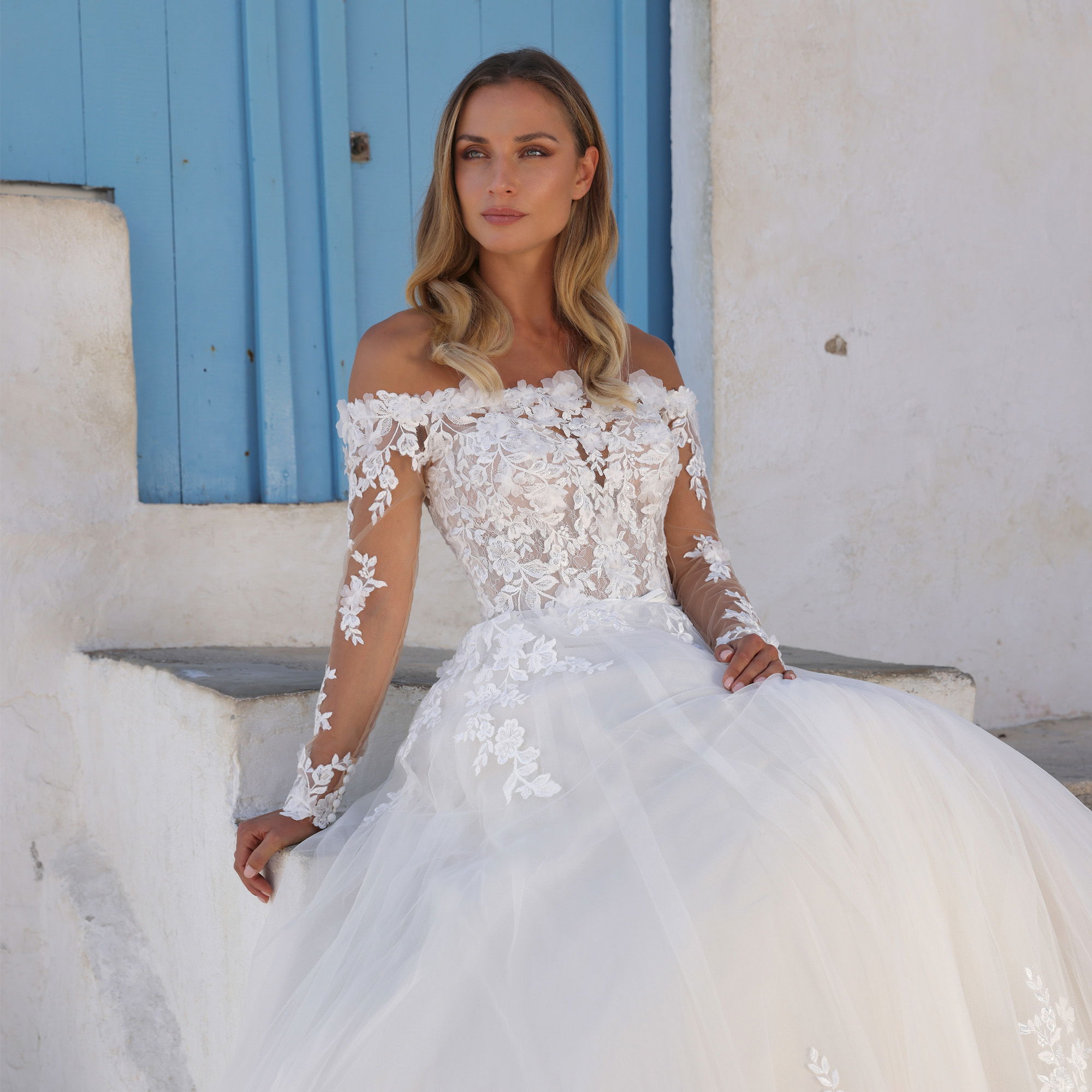 Verspieltes Brautkleid mit transparenten Ärmeln und blumigen Spitzendetails, ideal für eine Hochzeit in einer idyllischen, mediterranen Umgebung.