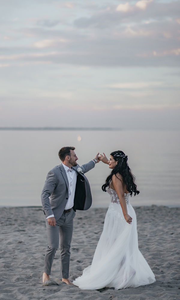 Ein frisch verheiratetes Paar tanzt am Ufer eines stillen Sees. Der Bräutigam im grauen Anzug hebt die Hand der Braut, die ein elegantes Hochzeitskleid trägt.