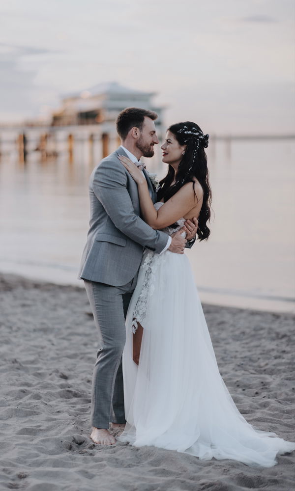 Ein verliebtes Brautpaar steht innig am Strand, der Bräutigam hält die Braut fest im Arm. Hinter ihnen erstreckt sich ein ruhiger See mit einem Steg.