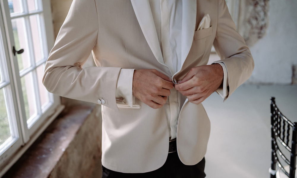Ein Mann im cremefarbenen Hochzeitsanzug knöpft sein Sakko. Das Licht durch die Fenster betont die feinen Details des Stoffes und die elegante Atmosphäre.