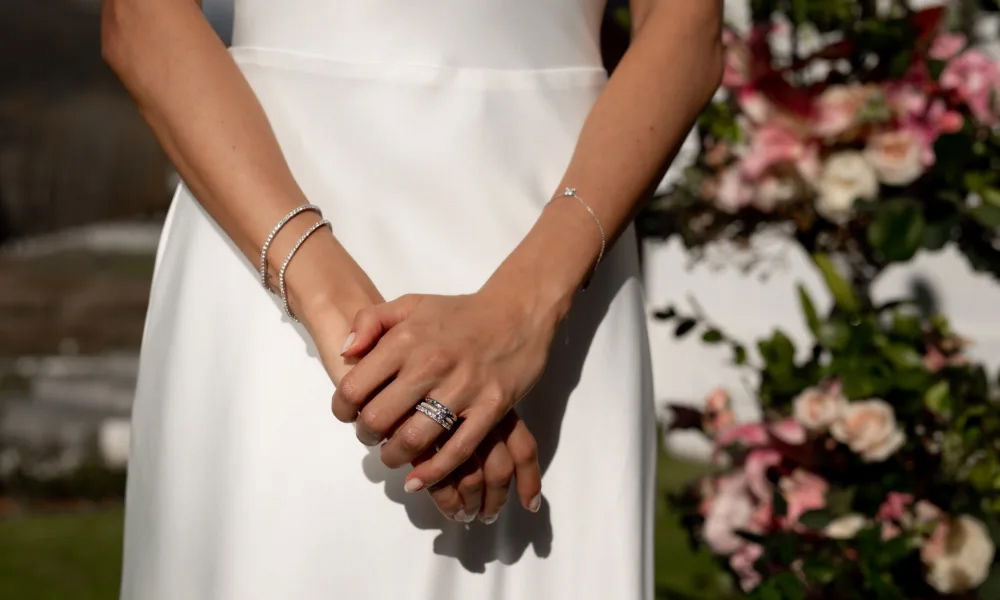 In einem eleganten Moment hält die Braut ihre Hände in einer harmonischen Pose, um ihren glänzenden Ehering und die dezenten Armbänder zu präsentieren. Die Diamanten auf dem Ring fangen das Sonnenlicht ein, während die blühende Natur im Hintergrund eine beruhigende Wirkung ausstrahlt.