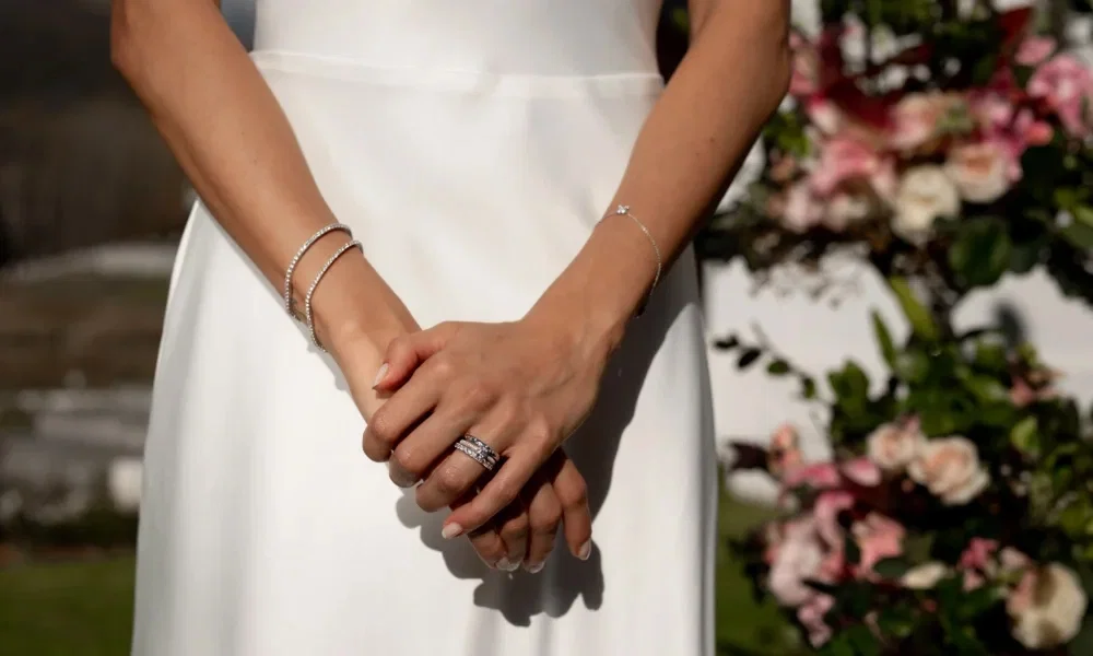 In einem eleganten Moment hält die Braut ihre Hände in einer harmonischen Pose, um ihren glänzenden Ehering und die dezenten Armbänder zu präsentieren. Die Diamanten auf dem Ring fangen das Sonnenlicht ein, während die blühende Natur im Hintergrund eine beruhigende Wirkung ausstrahlt.
