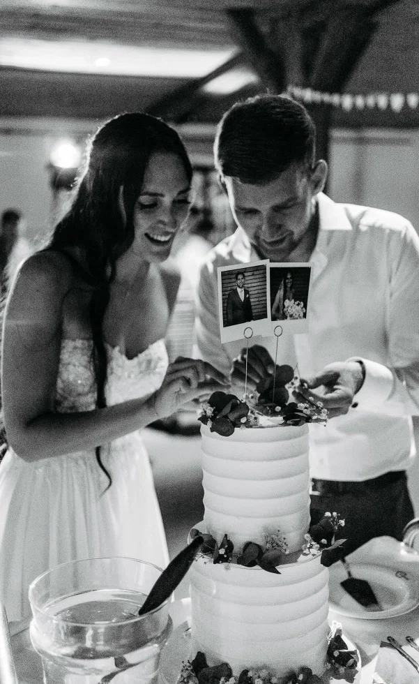 Brautpaar schneidet lachend die Hochzeitstorte an, dekoriert mit Blumen und persönlichen Polaroid-Fotos als Cake Topper in stimmungsvoller Schwarz-Weiß-Aufnahme