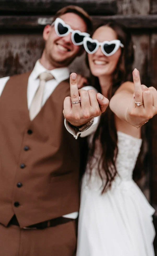 Lockeres Hochzeitsfoto: Braut und Bräutigam lachen, tragen verspielte Herzbrillen und präsentieren ihre Hände mit sichtbaren Trauringen.