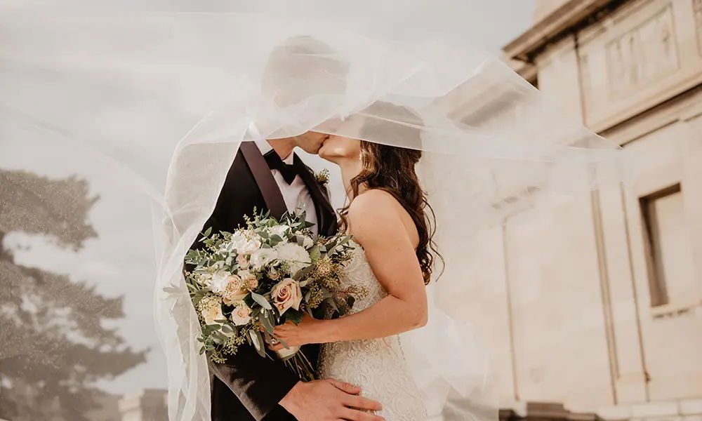 Ein inniger Kuss eines Brautpaares, eingehüllt in einen langen Schleier. Die Braut trägt ein weißes Spitzenkleid und hält einen großen Blumenstrauß, der Bräutigam trägt einen eleganten schwarzen Anzug.