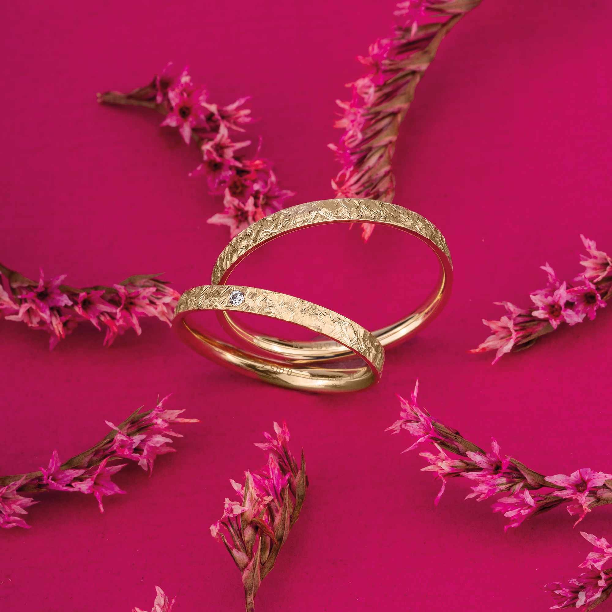 Stilvolle goldene Trauringe in einem romantischen Blumen-Setting auf einem leuchtend pinken Untergrund.