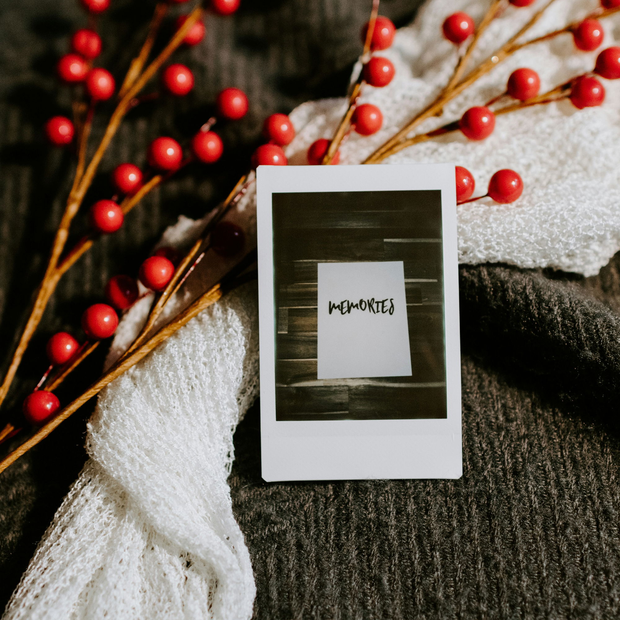 Detailaufnahme eines Polaroids mit "Memories", dekoriert mit roten Beerenzweigen und einem weißen Strickschal auf einer dunklen Stoffunterlage.