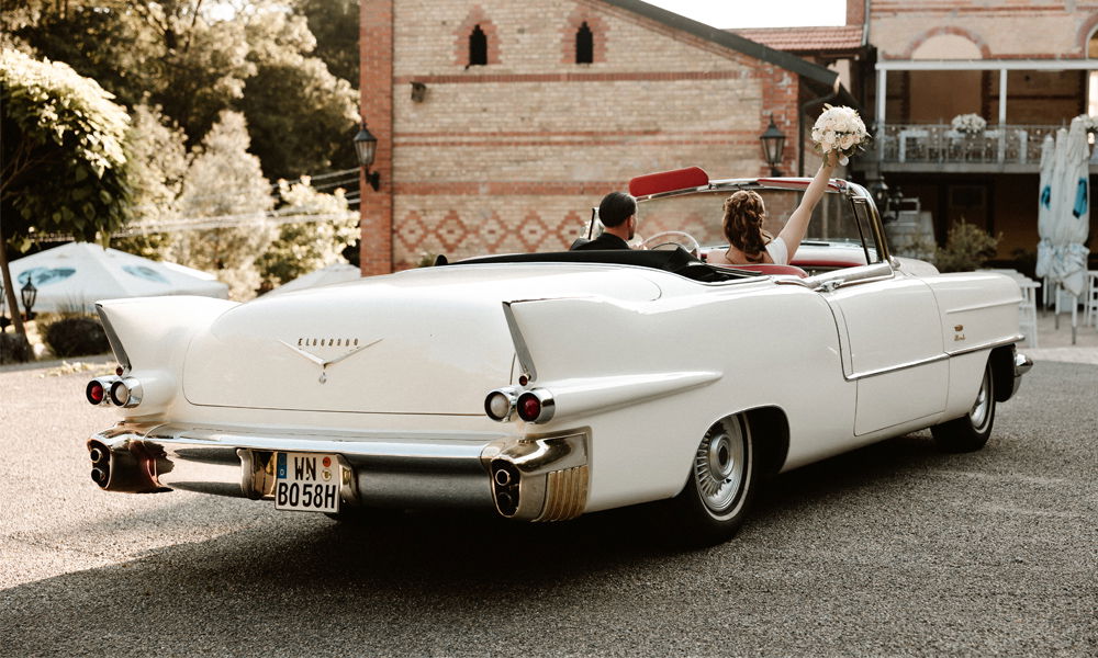 Rückansicht eines weißen Oldtimers mit offenem Verdeck, in dem ein Brautpaar sitzt. Die Braut hebt einen Brautstrauß, während das Fahrzeug auf einem idyllischen Gelände steht.