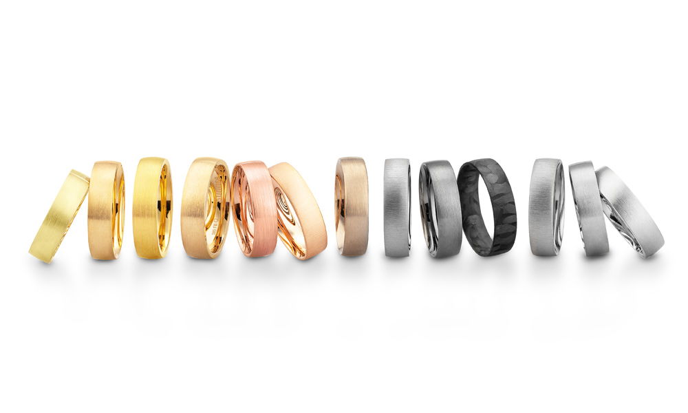 Moderne Eheringe in unterschiedlichen Metallfarben, von klassischen Goldtönen über Roségold bis hin zu zeitlosem Weißgold und außergewöhnlichem Schwarz. Die Designs variieren zwischen polierten und gebürsteten Oberflächen, teils mit markanter Struktur.