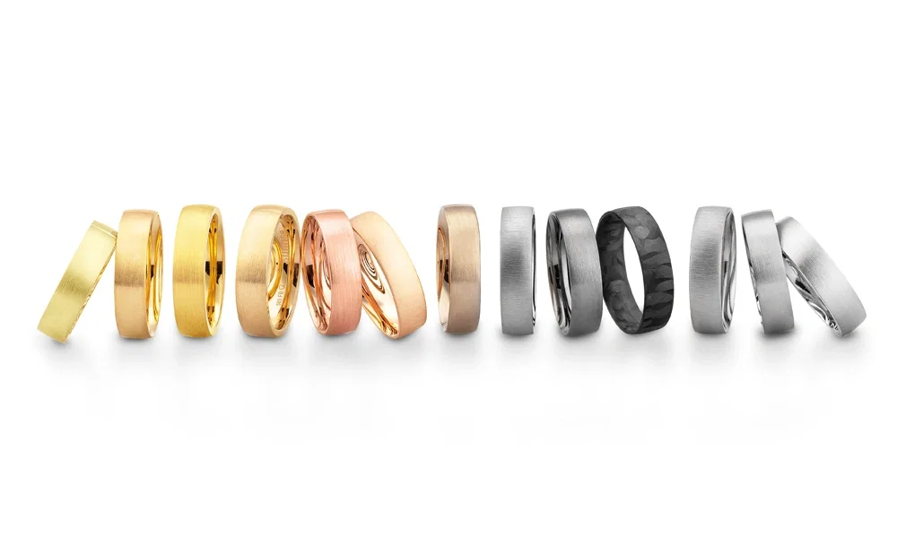 Moderne Eheringe in unterschiedlichen Metallfarben, von klassischen Goldtönen über Roségold bis hin zu zeitlosem Weißgold und außergewöhnlichem Schwarz. Die Designs variieren zwischen polierten und gebürsteten Oberflächen, teils mit markanter Struktur.
