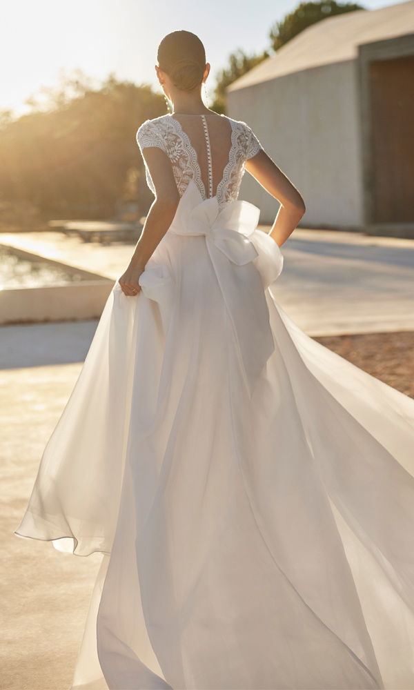 Eine stilvolle Braut in einem luftigen Hochzeitskleid mit offener Rückenpartie und großer Schleife, die elegant im Wind weht.