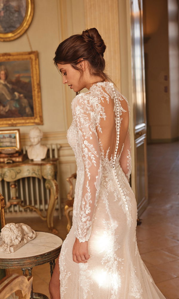 Eine stilvolle Braut in einem Hochzeitskleid mit filigraner Spitze, figurbetontem Schnitt und transparenten Ärmeln, perfekt für eine klassische Hochzeit.