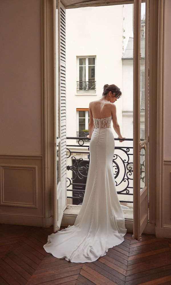 Eine Braut trägt ein modernes, enganliegendes Hochzeitskleid mit offener Rückenpartie und Knopfleiste, umgeben von klassischer Pariser Architektur.