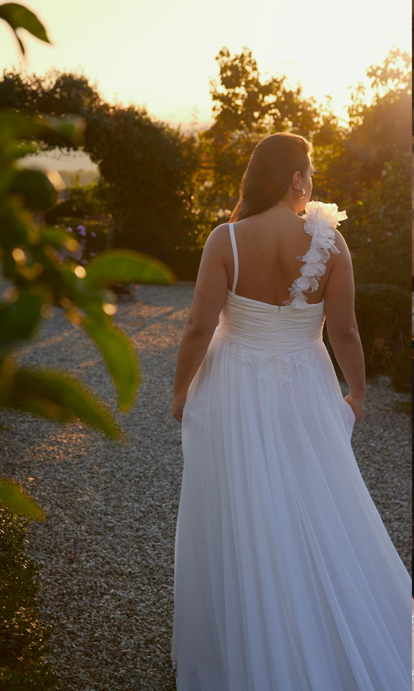 Die Braut schreitet in einem eleganten, fließenden Hochzeitskleid mit floralem Träger durch einen romantischen Garten im Abendlicht.