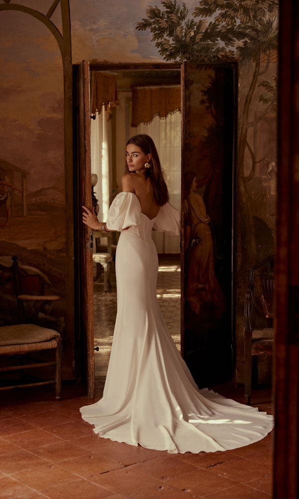 Eine elegante Braut in einem schmalen Kleid mit Schleppe und freien Schultern steht in einem kunstvoll gestalteten Raum mit offenen Türen.