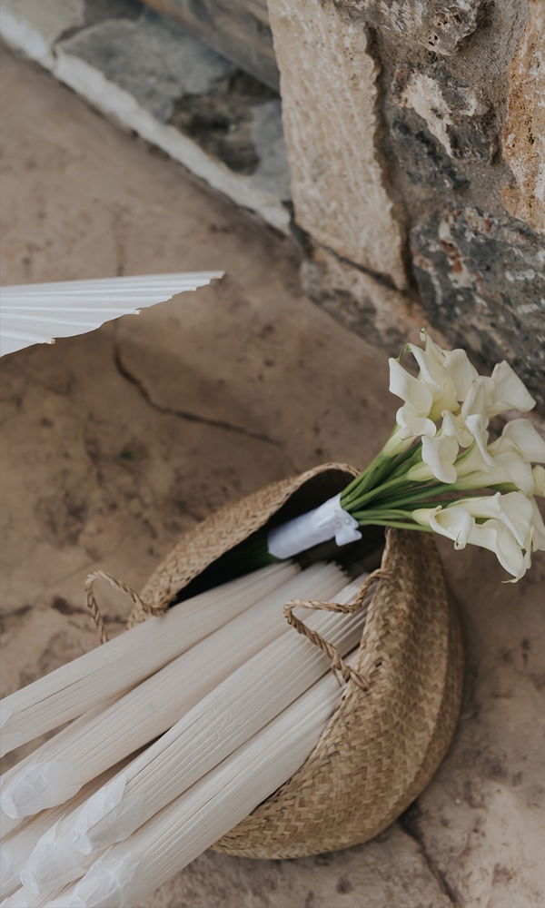 Natürliche Hochzeitsdetails: Ein schlichter Brautstrauß mit Calla-Lilien ruht in einer Korbtasche mit Sonnenschirmen für die Gäste.