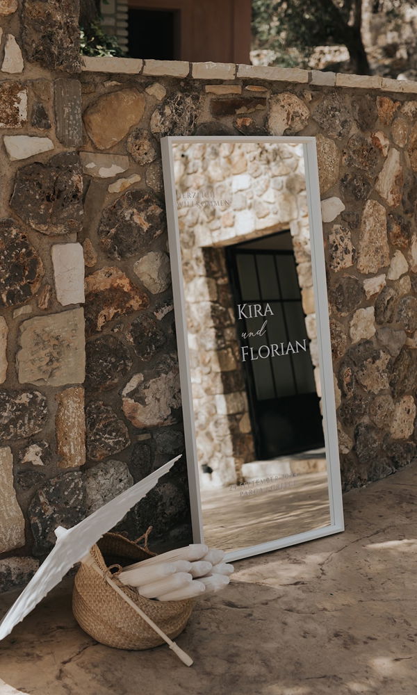 Modernes Willkommensschild mit den Namen des Brautpaares auf einem großen Spiegel, elegant in eine mediterrane Steinwand eingebettet.
