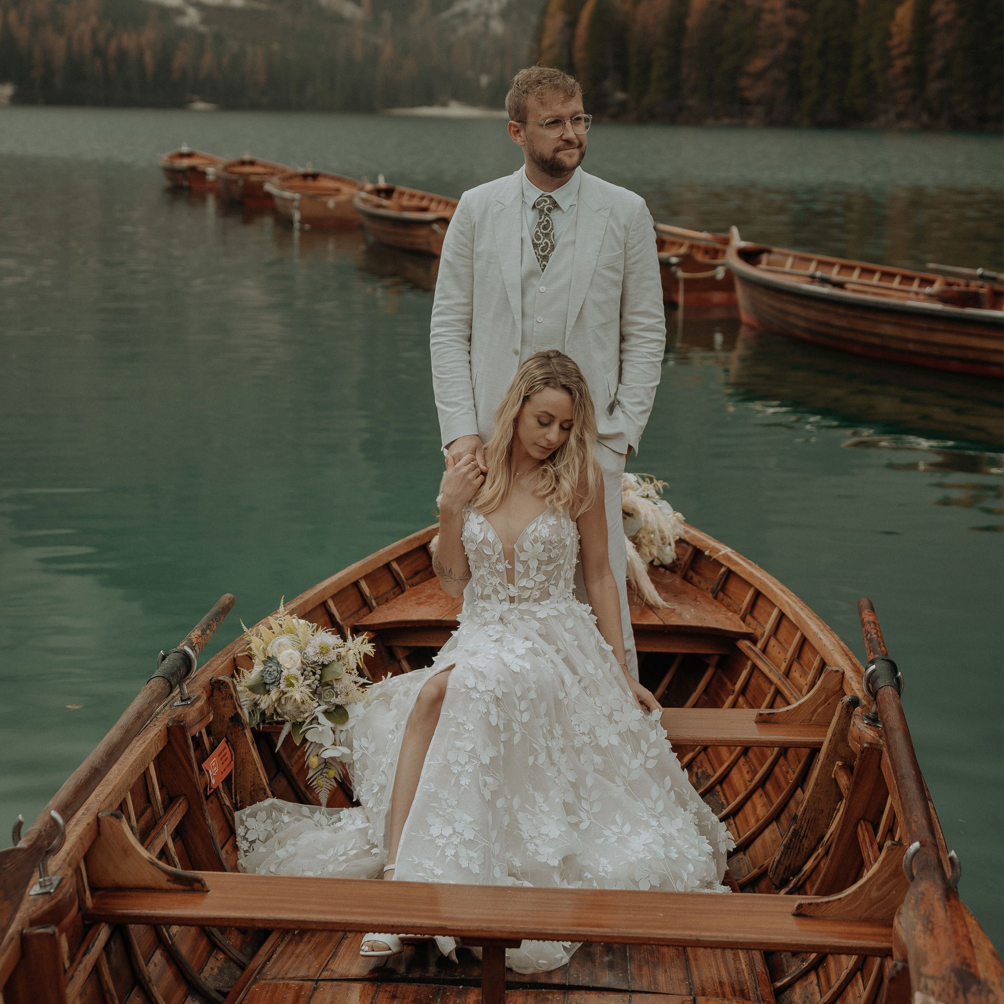 Ein frisch vermähltes Paar genießt eine Fahrt auf einem See. Die Braut sitzt anmutig mit ihrem langen Kleid, während der Bräutigam mit Brille und Anzug hinter ihr steht. Die traditionelle Holzboot-Kulisse verleiht dem Bild eine nostalgische Note.